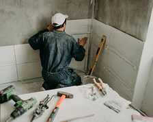 A man in blue work coveralls kneels in a bathroom corner to install large white tiles on the wall.